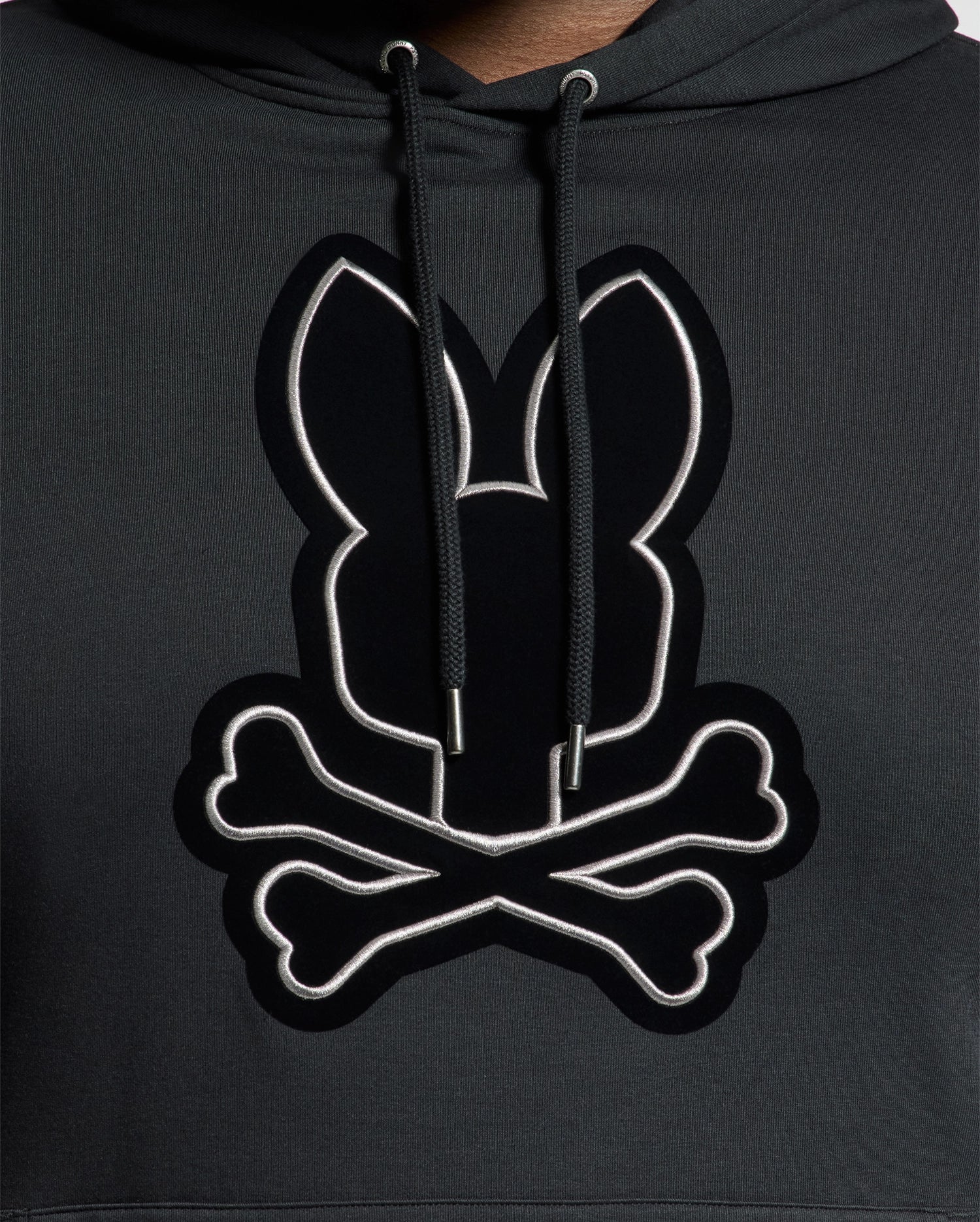 The Psycho Bunny BIG AND TALL ALEXANDER HOODIE - B9H154E200 is a black pullover hoodie featuring a stylized rabbit head with crossed bones beneath it. The design is outlined in white, providing striking contrast against the dark fabric. This comfortable hoodie is available in big and tall sizes for an ideal fit for everyone.