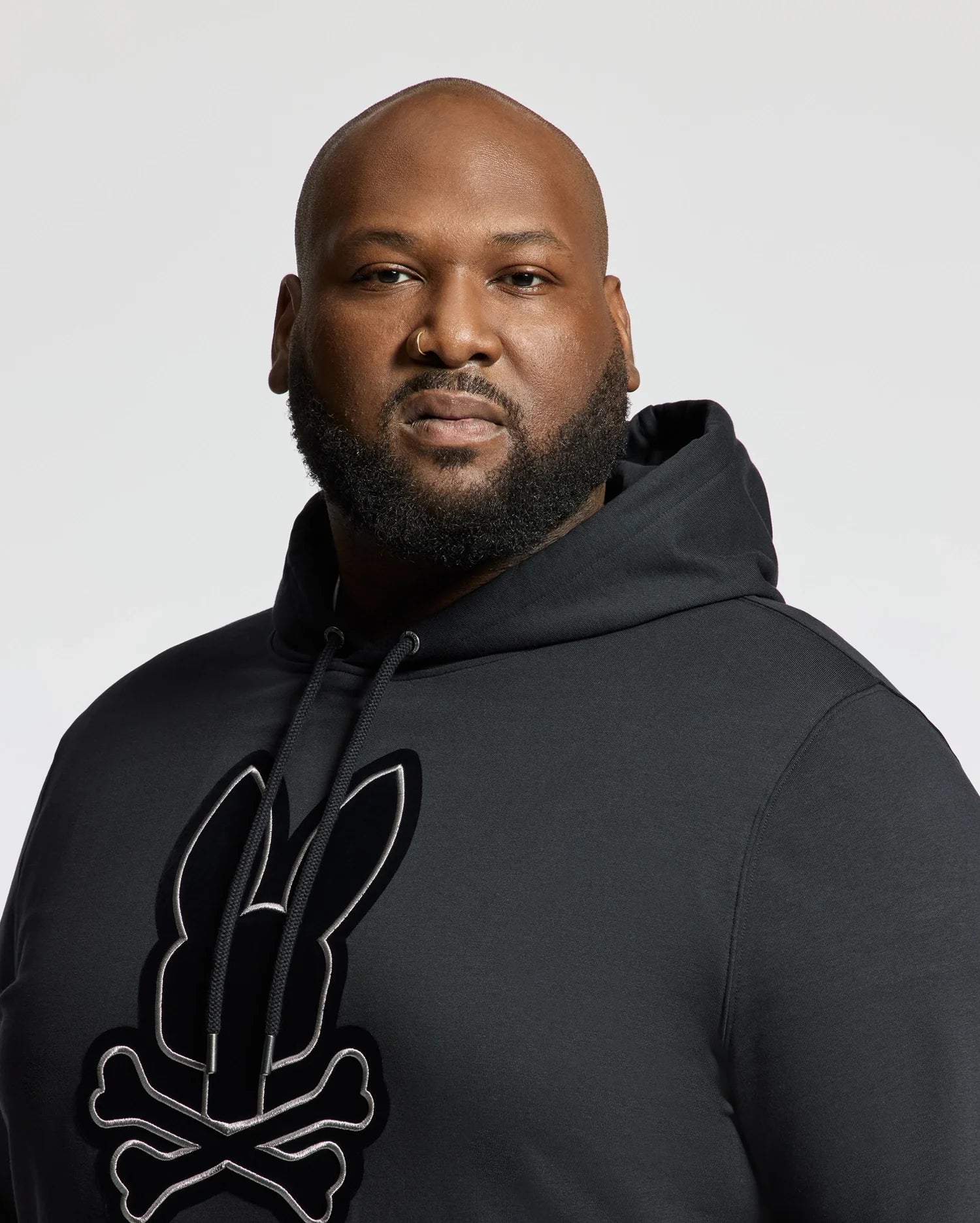A man with a beard is wearing the Psycho Bunny BIG AND TALL ALEXANDER HOODIE - B9H154E200, a comfortable black pullover hoodie adorned with a rabbit skeleton design, as he faces the camera against a plain, light-colored background.