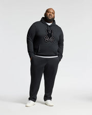 A man stands against a plain backdrop, wearing the Psycho Bunny BIG AND TALL ALEXANDER HOODIE - B9H154E200 in black, featuring a stylized graphic. He complements it with matching black sweatpants and white sneakers, achieving a casual and sporty look.