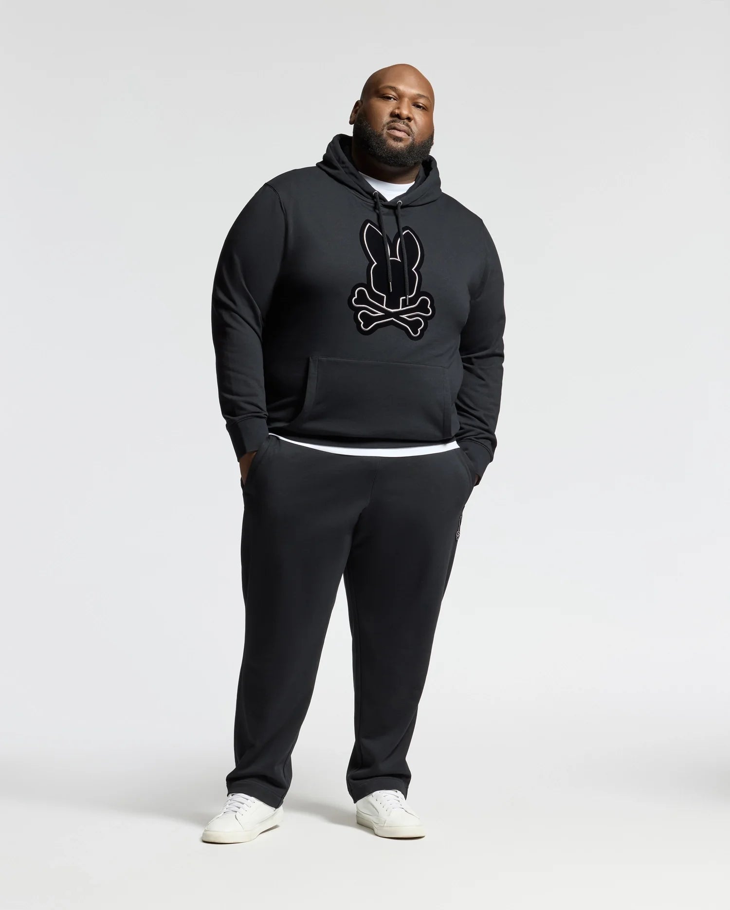A man stands against a plain backdrop, wearing the Psycho Bunny BIG AND TALL ALEXANDER HOODIE - B9H154E200 in black, featuring a stylized graphic. He complements it with matching black sweatpants and white sneakers, achieving a casual and sporty look.