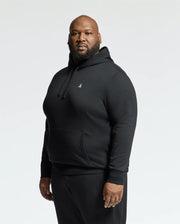 A person dressed in a Psycho Bunny Men's Big and Tall Levy Essential Fleece Hoodie - B9H859D200, featuring a kangaroo pocket, pairs it with black pants as they stand against a plain white background, gazing at the camera. Made from 100% cotton, this outfit seamlessly merges comfort with style.