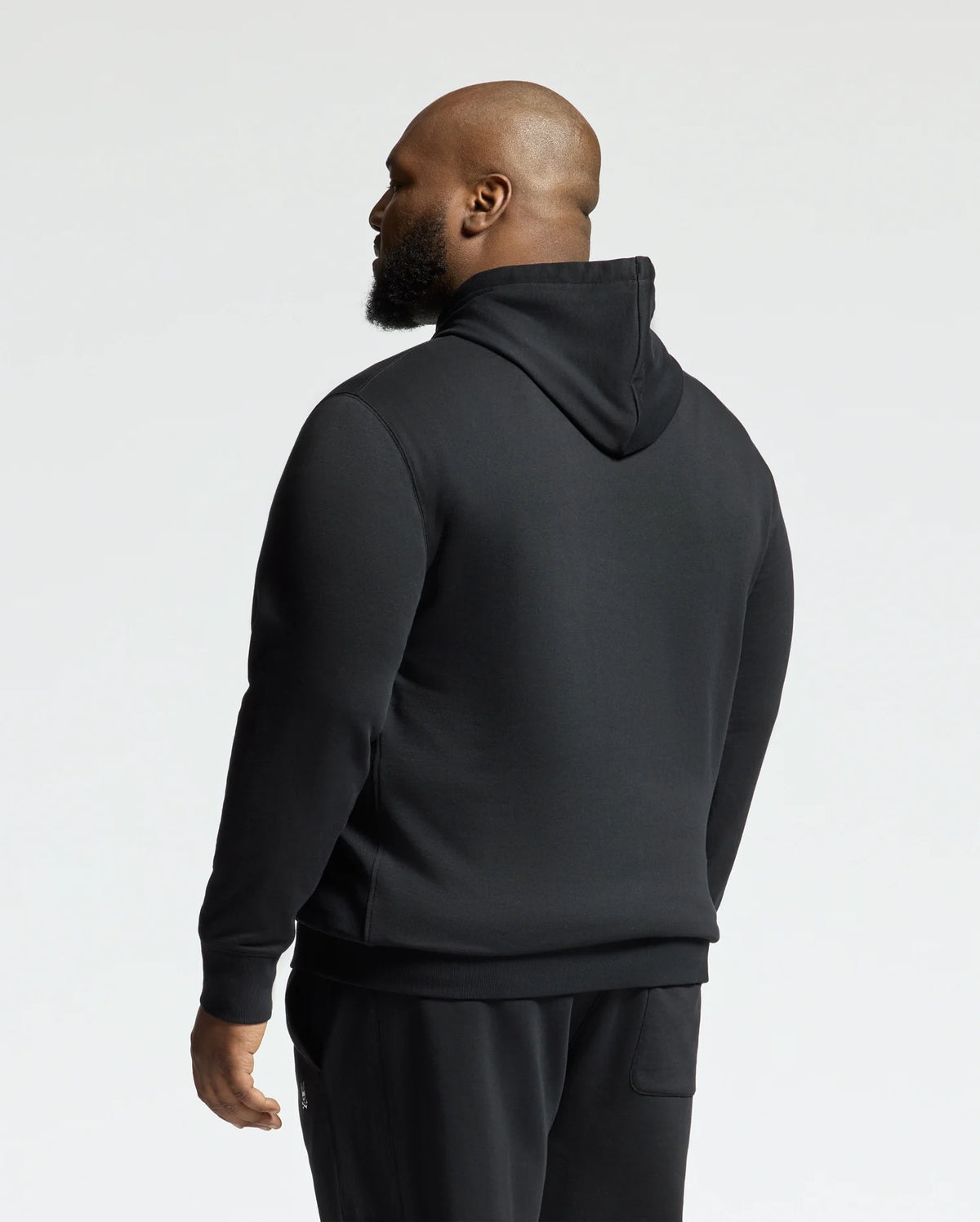 A person with a beard is wearing a black Psycho Bunny MENS BIG AND TALL LEVY ESSENTIAL FLEECE HOODIE - B9H859D200 and black pants, facing away from the camera. The 100% cotton hoodie includes a kangaroo pocket, combining style and functionality. The background is plain and light-colored.