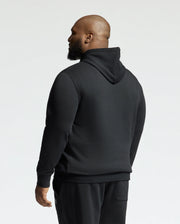 A person with a beard is wearing a black Psycho Bunny MENS BIG AND TALL LEVY ESSENTIAL FLEECE HOODIE - B9H859D200 and black pants, facing away from the camera. The 100% cotton hoodie includes a kangaroo pocket, combining style and functionality. The background is plain and light-colored.