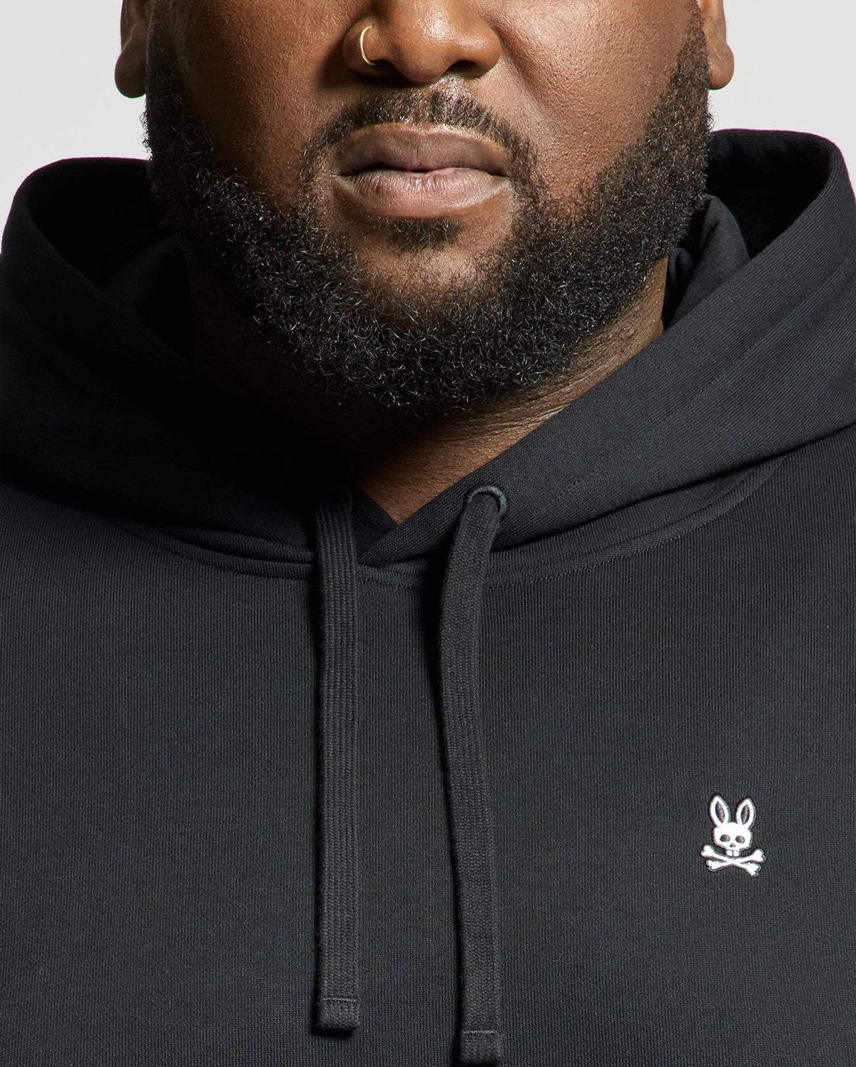 A person sporting a black Psycho Bunny MENS BIG AND TALL LEVY ESSENTIAL FLEECE HOODIE - B9H859D200, adorned with a small white bunny and crossbones logo. Made from 100% cotton, the hoodie includes a kangaroo pocket. The individual, who has a beard and nose ring, is set against a plain background that highlights the upper half of the hoodie and their face.