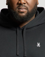 A person sporting a black Psycho Bunny MENS BIG AND TALL LEVY ESSENTIAL FLEECE HOODIE - B9H859D200, adorned with a small white bunny and crossbones logo. Made from 100% cotton, the hoodie includes a kangaroo pocket. The individual, who has a beard and nose ring, is set against a plain background that highlights the upper half of the hoodie and their face.
