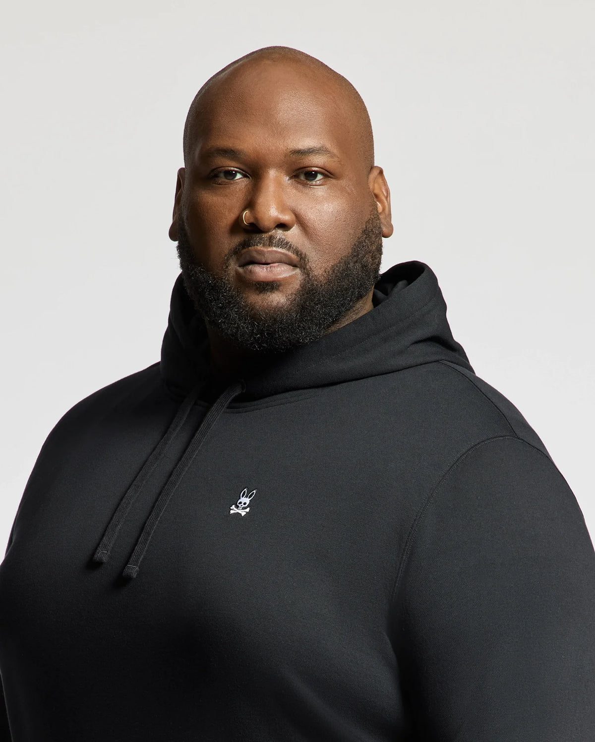 A man is confidently looking at the camera wearing a Psycho Bunny Men's Big and Tall Levy Essential Fleece Hoodie, B9H859D200, which features a small white logo on its chest. This black sweatshirt, made from 100% cotton, includes a cozy kangaroo pocket. The background is a plain off-white color.
