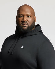 A man is confidently looking at the camera wearing a Psycho Bunny Men's Big and Tall Levy Essential Fleece Hoodie, B9H859D200, which features a small white logo on its chest. This black sweatshirt, made from 100% cotton, includes a cozy kangaroo pocket. The background is a plain off-white color.