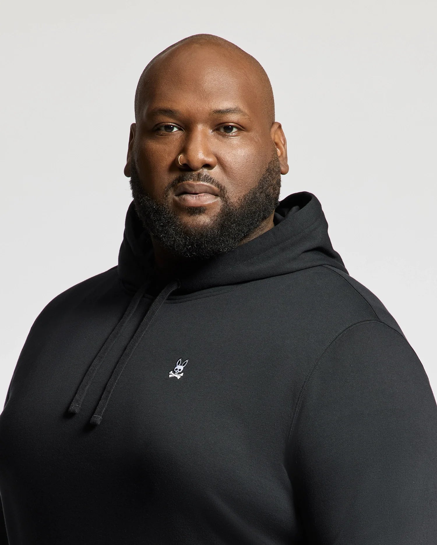 A man is confidently looking at the camera wearing a Psycho Bunny Men's Big and Tall Levy Essential Fleece Hoodie, B9H859D200, which features a small white logo on its chest. This black sweatshirt, made from 100% cotton, includes a cozy kangaroo pocket. The background is a plain off-white color.