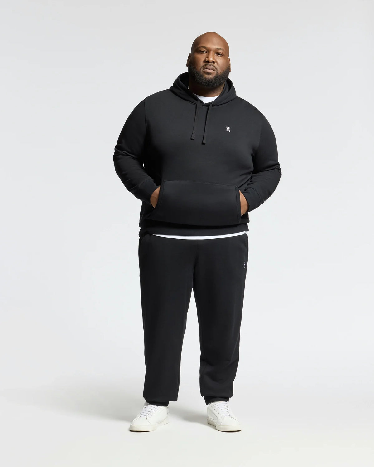A man dressed in a black "Mens Big and Tall Levy Essential Fleece Hoodie - B9H859D200" by Psycho Bunny and black sweatpants poses against a plain white background. Made from 100% cotton, the hoodie includes a convenient kangaroo pocket where he places his hands. His outfit is rounded off with white sneakers.