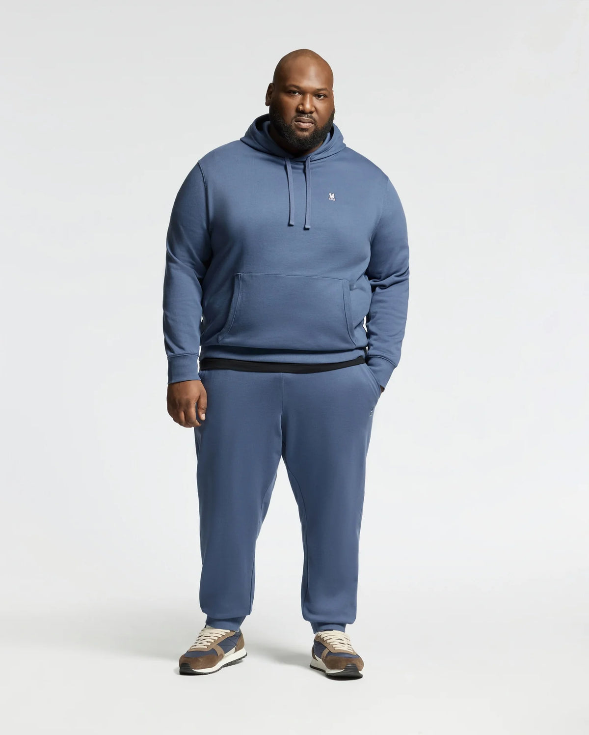 A man faces forward in a blue Psycho Bunny Mens Big and Tall Levy Essential Fleece Hoodie (B9H859D200), paired with matching sweatpants and brown sneakers, set against a plain light background.