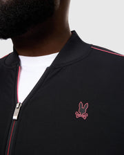 Close-up of a person wearing the Psycho Bunny BIG AND TALL TRAVIS TRACK JACKET (B9J142D200), a black cotton-blend piece with a pink rabbit and crossbones silicone logo on the chest, featuring a metal zipper pull and red piping along the seams, layered over a white shirt.