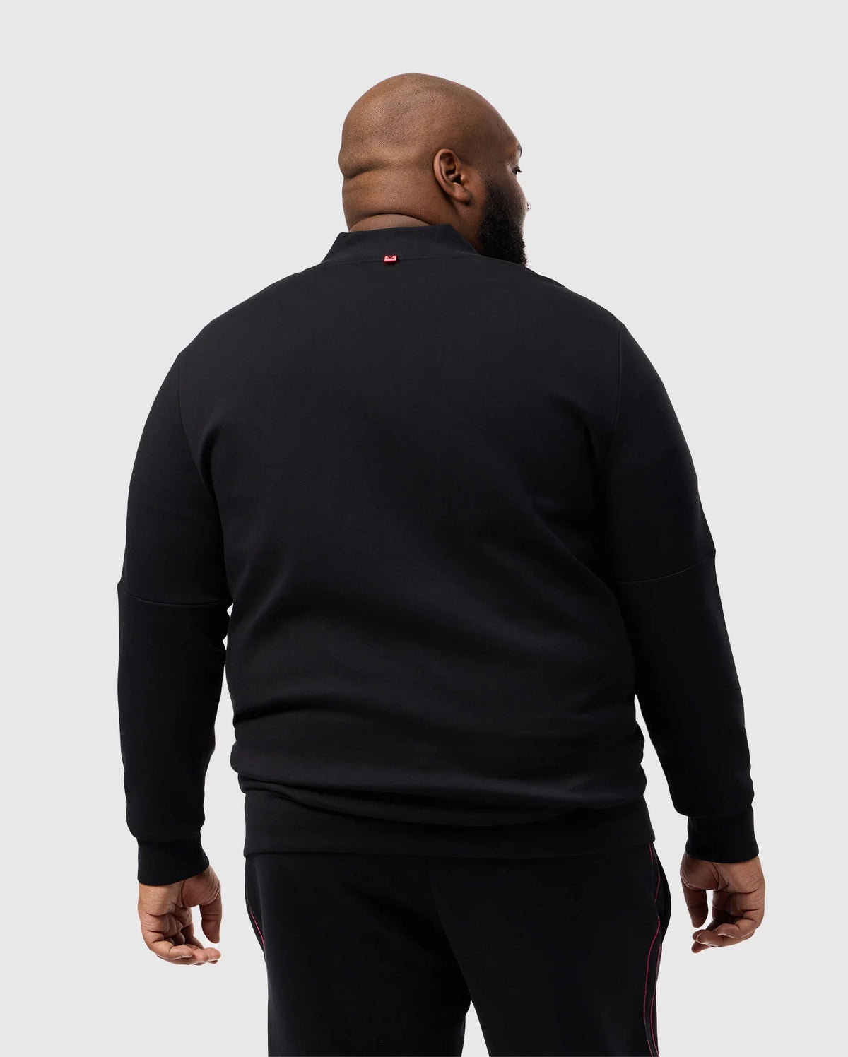 A man stands facing away in a Psycho Bunny BIG AND TALL TRAVIS TRACK JACKET, B9J142D200, against a light gray backdrop. The black cotton-blend jacket features a small red silicone heat-transfer logo near the collar for added style.