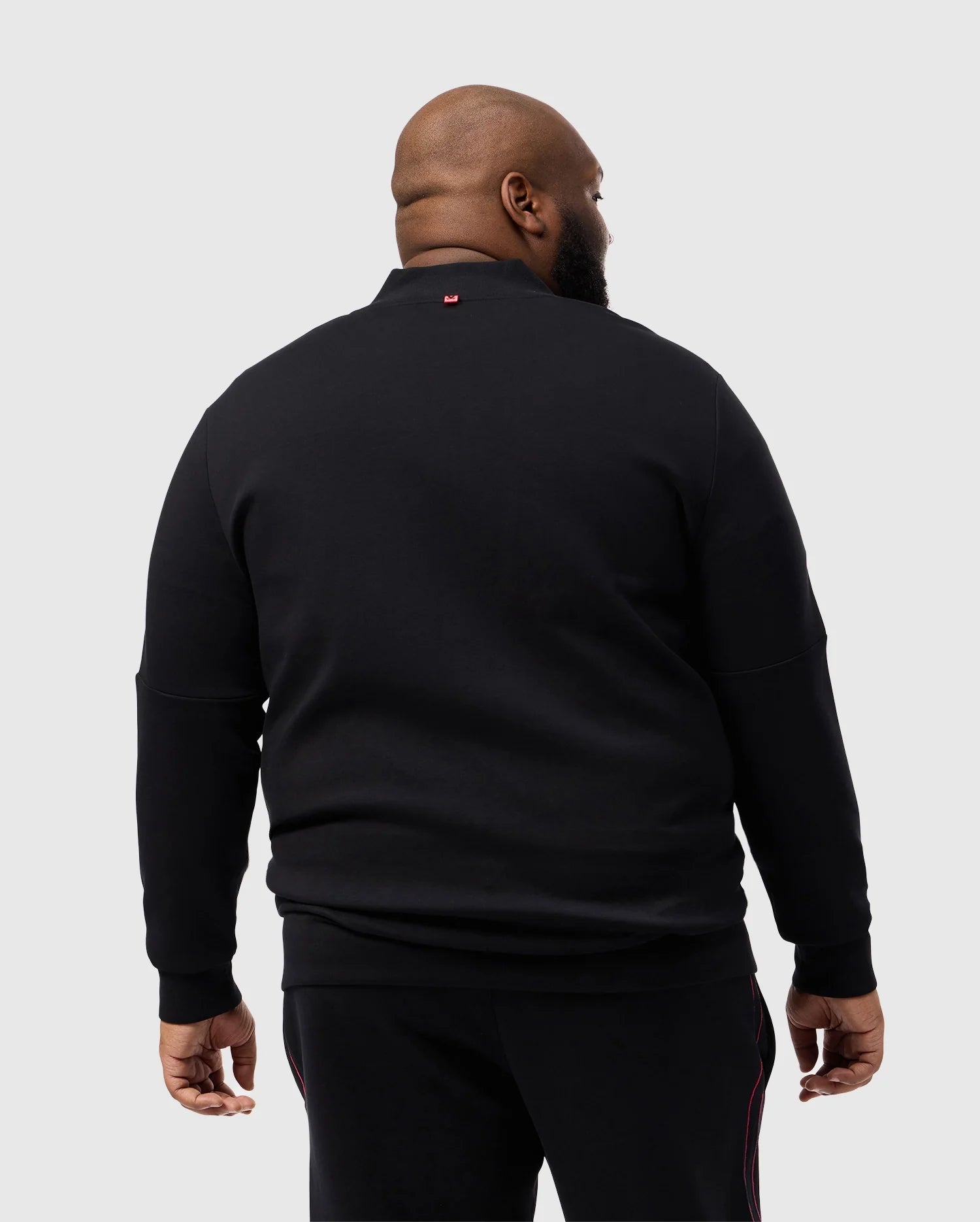 A man stands facing away in a Psycho Bunny BIG AND TALL TRAVIS TRACK JACKET, B9J142D200, against a light gray backdrop. The black cotton-blend jacket features a small red silicone heat-transfer logo near the collar for added style.