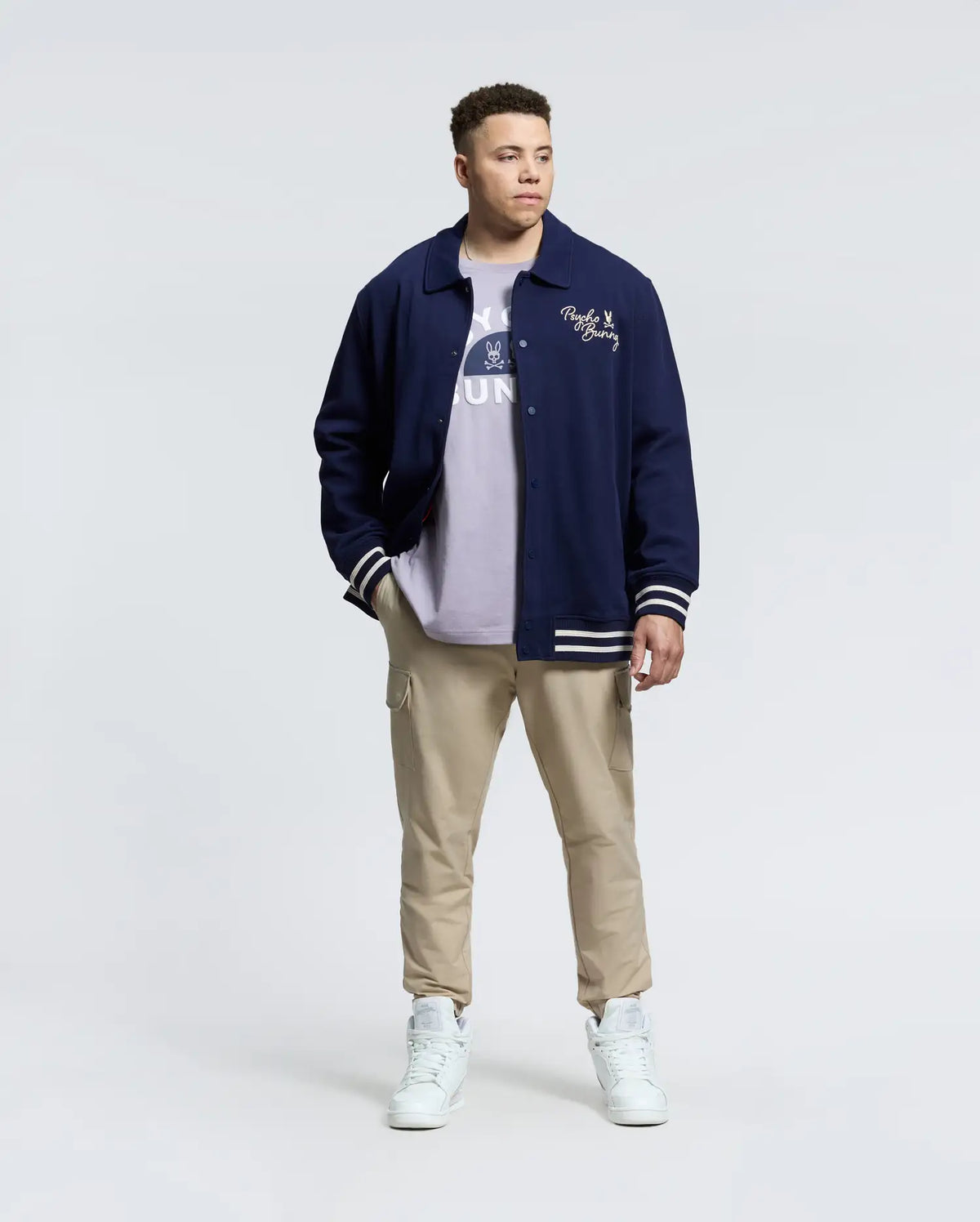 A person in a Psycho Bunny MENS BIG AND TALL DESERT COACH BOMBER JACKET - B9J428F200 stands against a plain background, wearing a light shirt, beige cargo pants, and white high-tops. They have short hair and hands in their pockets.