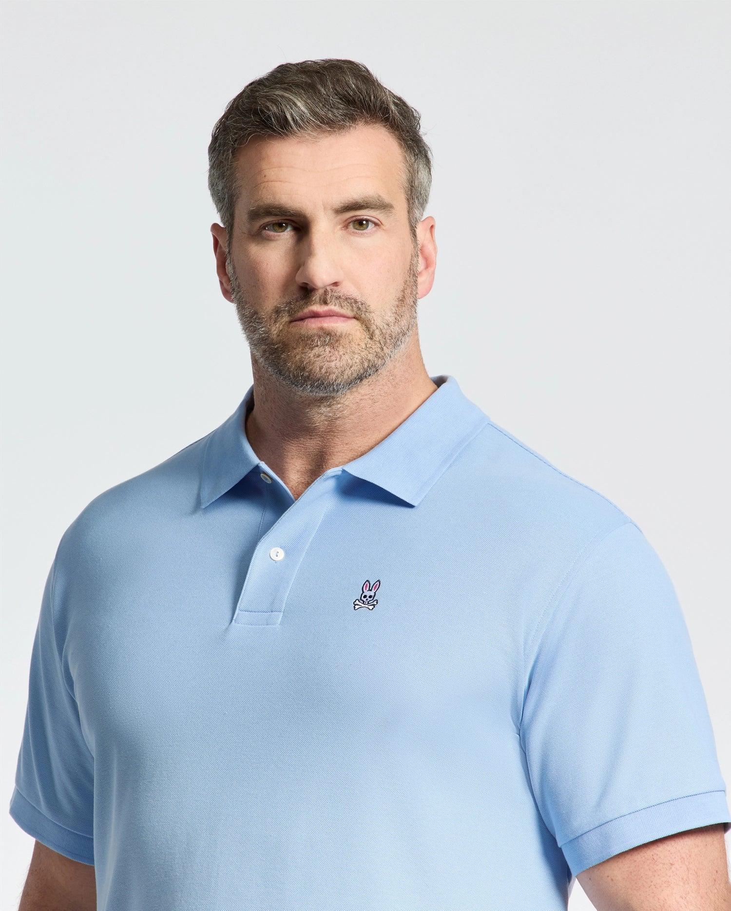 A man with short hair and a beard wears a Psycho Bunny BIG AND TALL CLASSIC PIQUE POLO - B9K001CRPC in light blue, featuring an embroidered logo on the chest. He stands against a plain white background, looking at the camera.