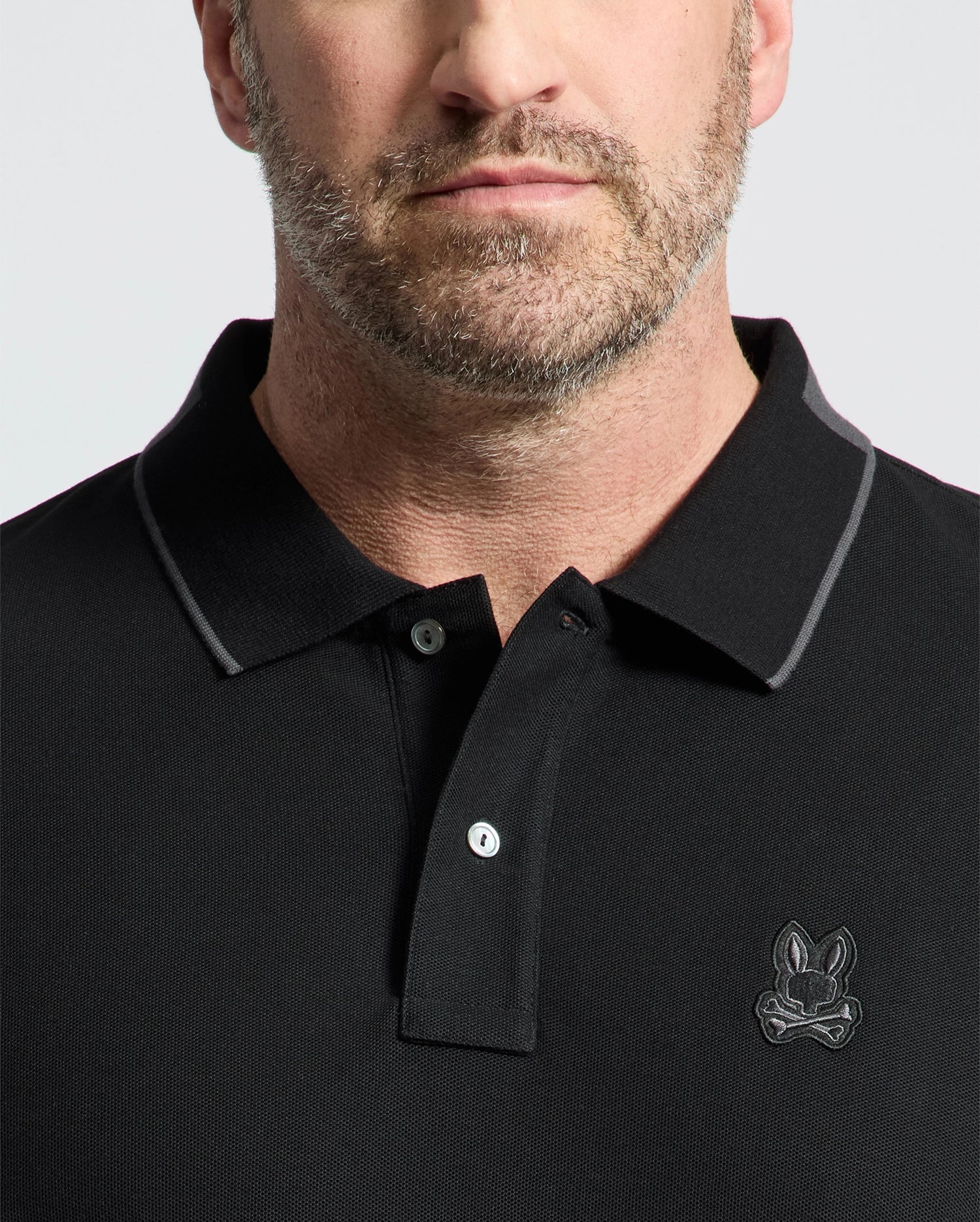 A man wears a black New Castle pique polo by Psycho Bunny, featuring a bunny logo on the chest. Made from 100% Pima cotton, it includes two buttons and luxury finishes like a subtle gray line around the collar. The background is plain light gray.