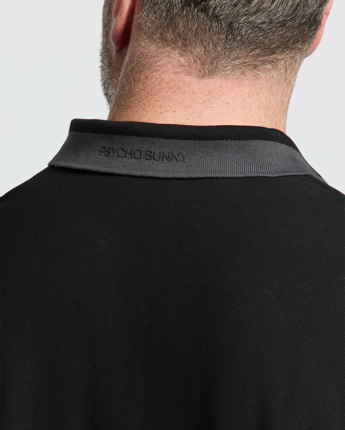 Close-up of a person's head and neck in a black MENS BIG AND TALL NEW CASTLE PIQUE POLO - B9K487F200 by Psycho Bunny. The collar has "Psycho Bunny" embroidered in black, highlighting the luxury. Made from 100% Pima cotton, it stands out against a plain white background.