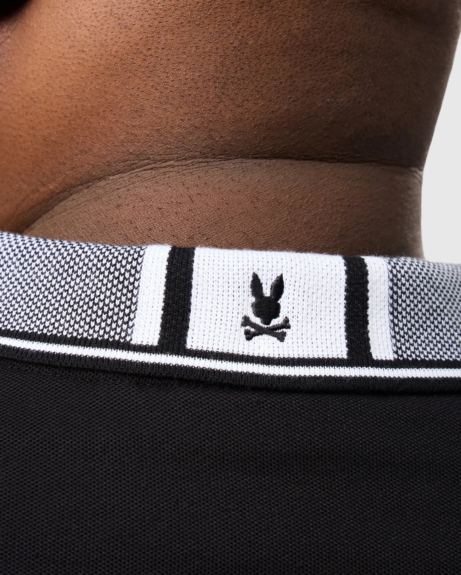 A close-up of a neckline shows a person wearing the BIG AND TALL MALTON PIQUE POLO SHIRT by Psycho Bunny, featuring a ribbed collar with a small black and white bunny logo above crossed bones.