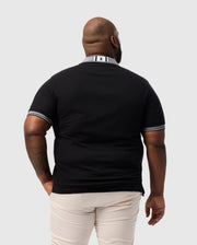 A person stands with their back to the camera in a Psycho Bunny Big and Tall Malton Pique Polo Shirt, featuring white-trimmed sleeves, made from soft Pima cotton, complemented by light-colored pants. The collar is subtly adorned with the Psycho Bunny logo against a plain gray backdrop.