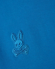 The blue BIG AND TALL MALTON PIQUE POLO SHIRT by Psycho Bunny features an embroidered rabbit skull and crossbones design with rabbit ears above crossed bones, set on a diamond pique pattern for added texture.