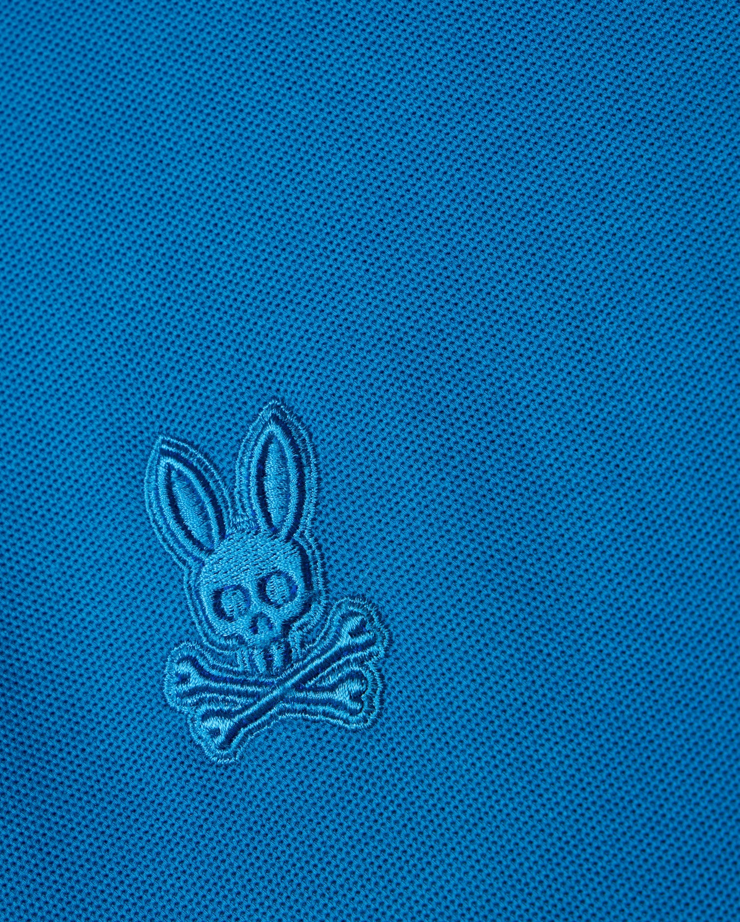 The blue BIG AND TALL MALTON PIQUE POLO SHIRT by Psycho Bunny features an embroidered rabbit skull and crossbones design with rabbit ears above crossed bones, set on a diamond pique pattern for added texture.