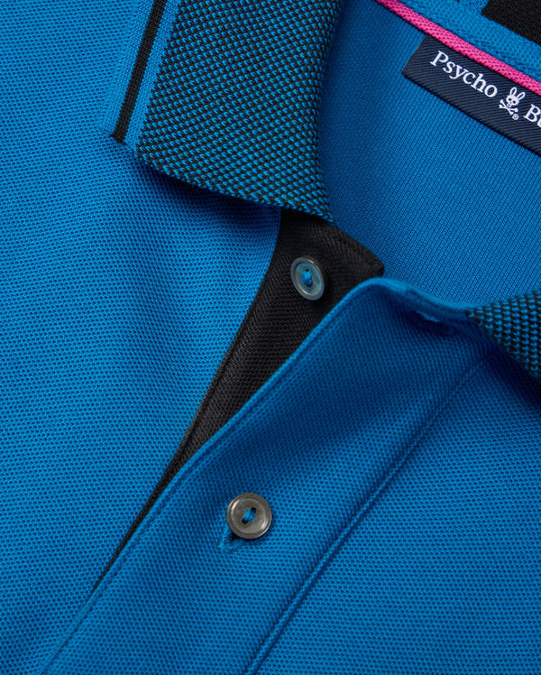 Close-up of a Psycho Bunny BIG AND TALL MALTON PIQUE POLO SHIRT - B9K950D200 in blue, featuring a textured collar with a black and blue diamond pique pattern, visible buttons on the placket, and an inside label showcasing its luxurious pima cotton craftsmanship.