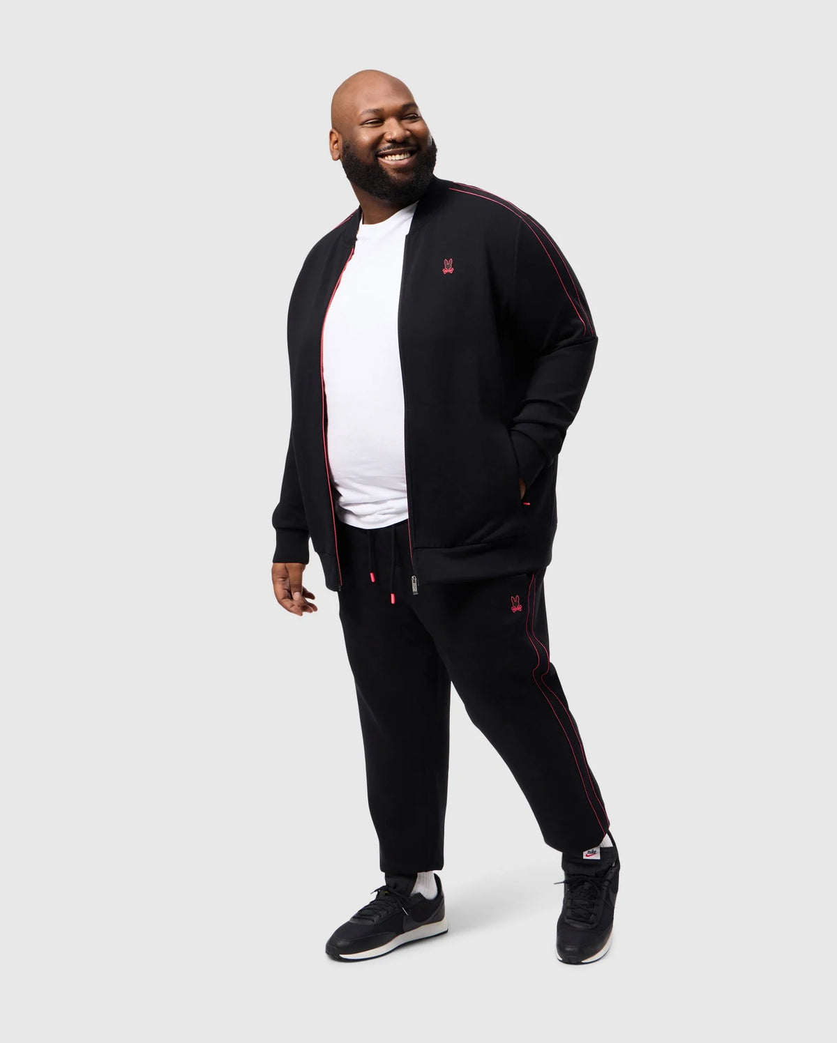 A man beams, wearing Psycho Bunny's BIG AND TALL TRAVIS TRACK PANT - B9P143D200, an elastic black tracksuit with red piping, and a white T-shirt. He pairs it with black sneakers, set against a plain white background, looking sideways with one hand in his pocket.