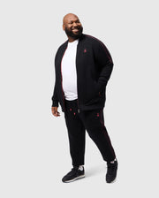 A man beams, wearing Psycho Bunny's BIG AND TALL TRAVIS TRACK PANT - B9P143D200, an elastic black tracksuit with red piping, and a white T-shirt. He pairs it with black sneakers, set against a plain white background, looking sideways with one hand in his pocket.