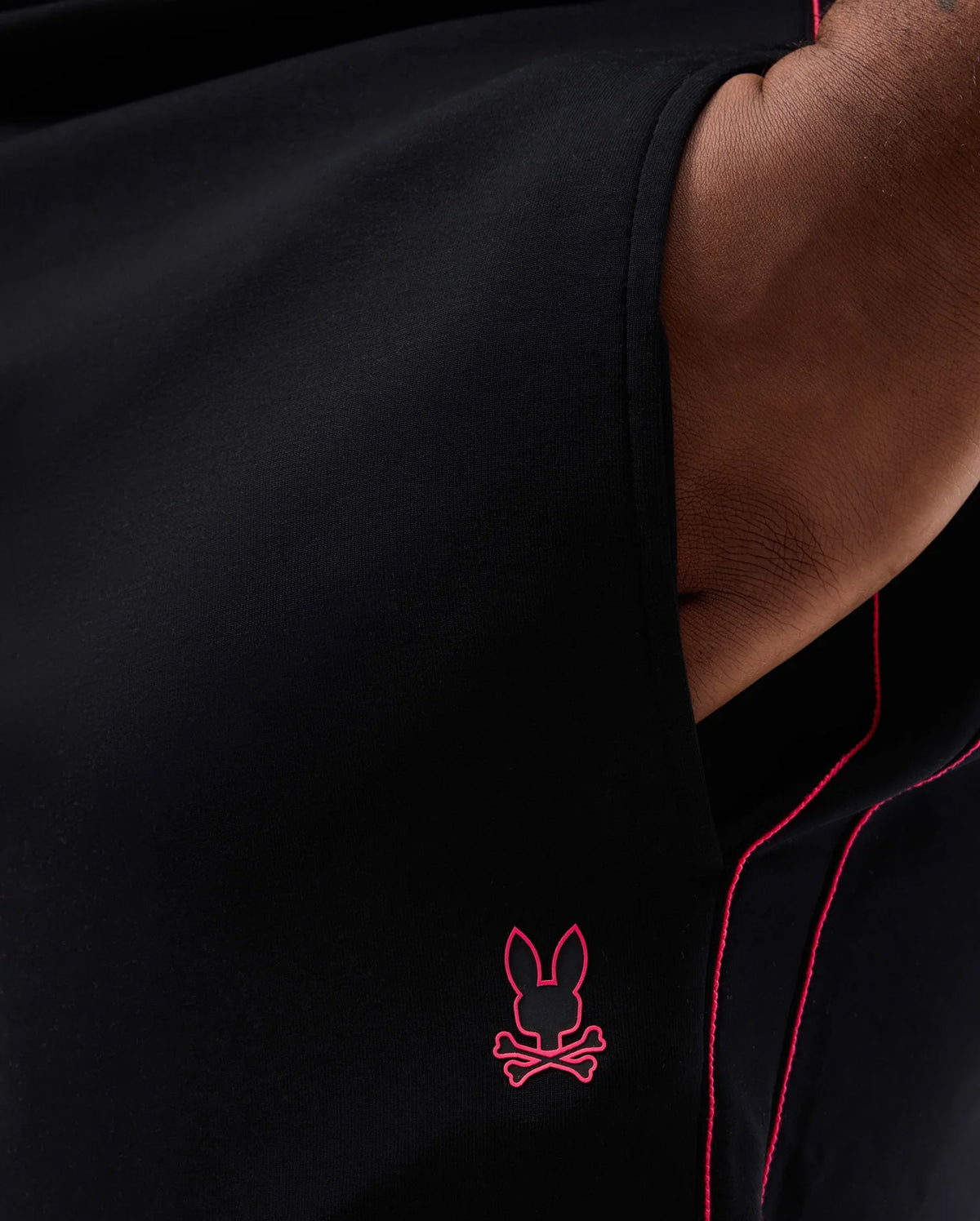 Close-up of a person wearing black clothing similar to the Psycho Bunny BIG AND TALL TRAVIS TRACK PANT - B9P143D200, featuring a small pink bunny with crossed bones emblem near the bottom, pink stitching along the edges, and an elastic waistband for added stretch.