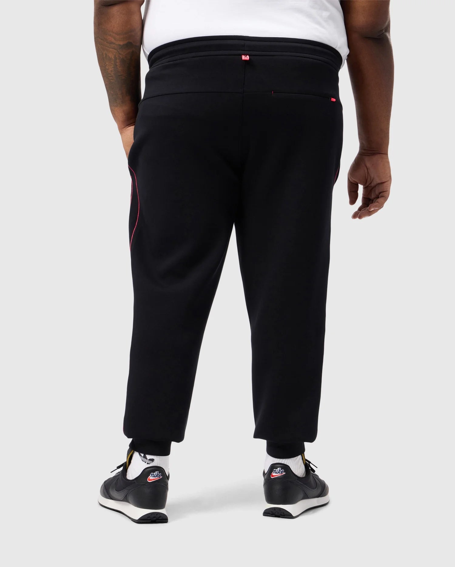 A person is standing with their back turned, wearing Psycho Bunny's BIG AND TALL TRAVIS TRACK PANT - B9P143D200 in black with red accents and black sneakers featuring white soles. The stretch fabric offers ease of movement against a plain, light-colored backdrop.