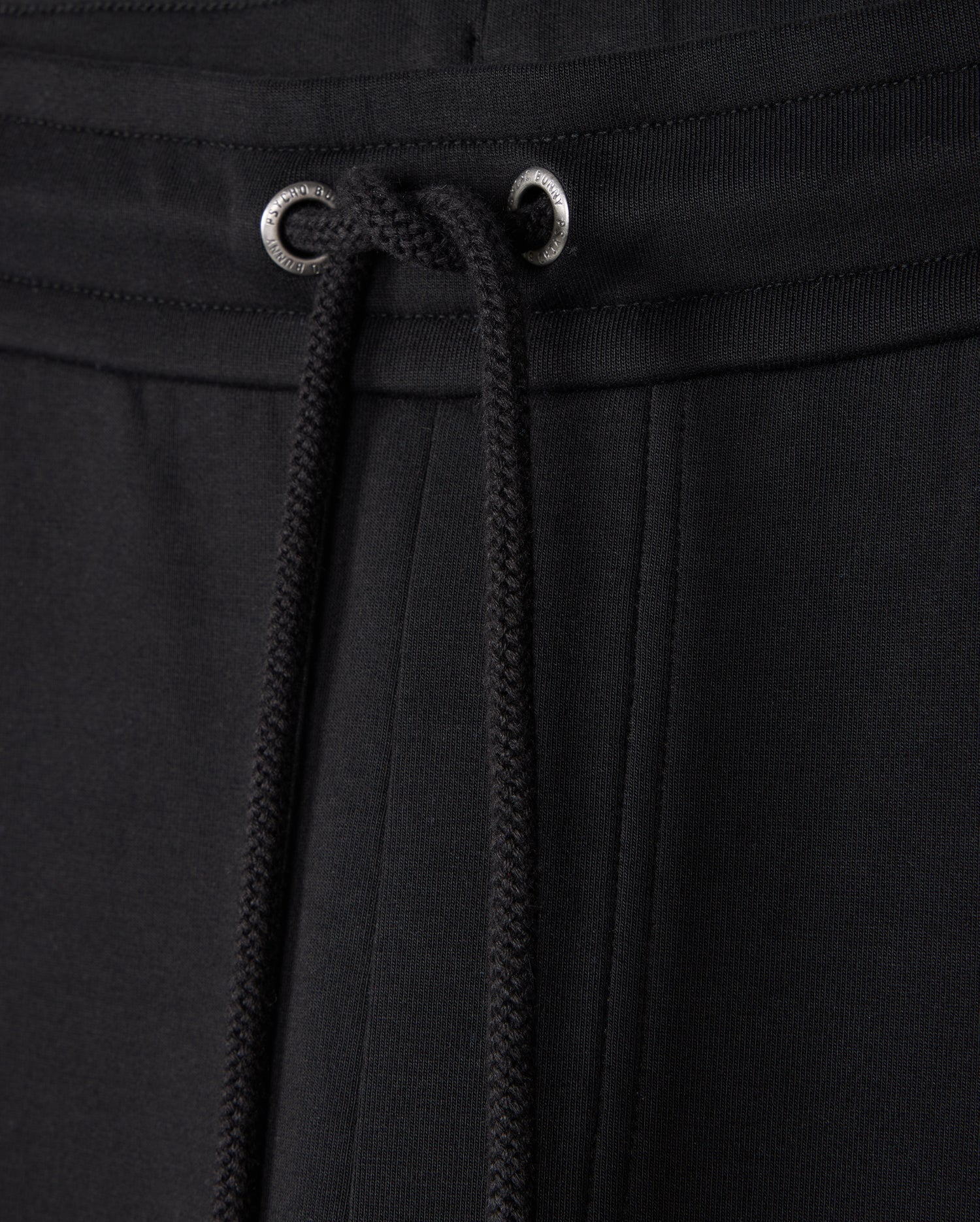 Close-up of BIG AND TALL Travis track pants by Psycho Bunny, featuring black fabric with metal eyelets. The soft, textured material and elastic drawstring waist ensure a casual, comfortable fit, perfect for leisurewear.