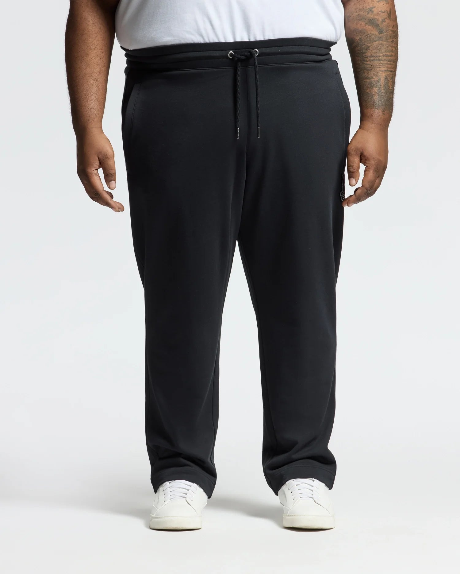 Someone in comfortable black Psycho Bunny BIG AND TALL ALEXANDER SWEATPANT - B9P155E200 and white sneakers stands against a white background. The joggers have a drawstring elastic waistband, and the person's left arm displays a tattoo.