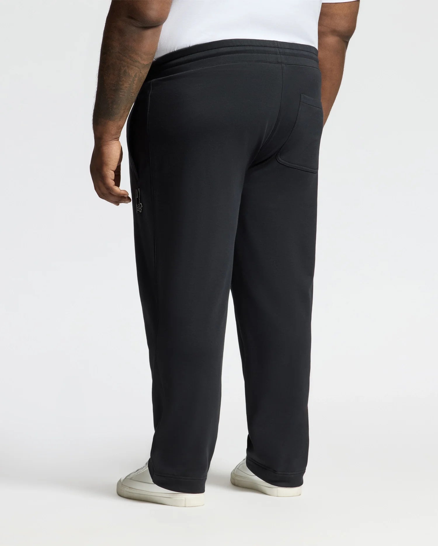 A person wearing Psycho Bunny's BIG AND TALL ALEXANDER SWEATPANT (B9P155E200) in black and white sneakers stands facing away, captured from the waist down. The scene is well-lit with a plain background, emphasizing their comfortable style in a white shirt.