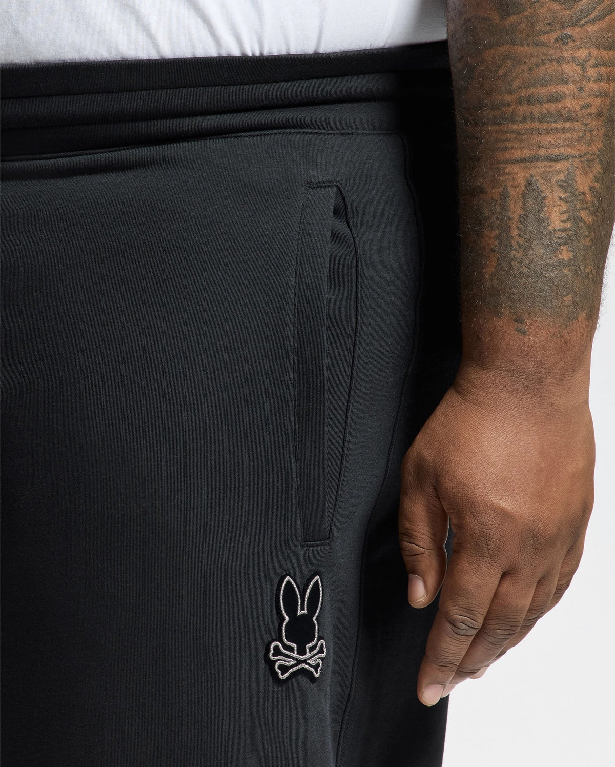 A person sporting Psycho Bunny's black BIG AND TALL ALEXANDER SWEATPANT (B9P155E200) with a bunny and crossbones logo on the pocket flaunts an intricately tattooed right arm. They're comfortably paired with a white shirt visible above the waistband.