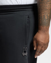 A person sporting Psycho Bunny's black BIG AND TALL ALEXANDER SWEATPANT (B9P155E200) with a bunny and crossbones logo on the pocket flaunts an intricately tattooed right arm. They're comfortably paired with a white shirt visible above the waistband.