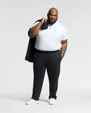 A man with a neutral expression and beard stands against a plain background, holding a jacket over his shoulder. He is dressed in a white t-shirt, BIG AND TALL ALEXANDER SWEATPANT - B9P155E200 by Psycho Bunny, and white sneakers.