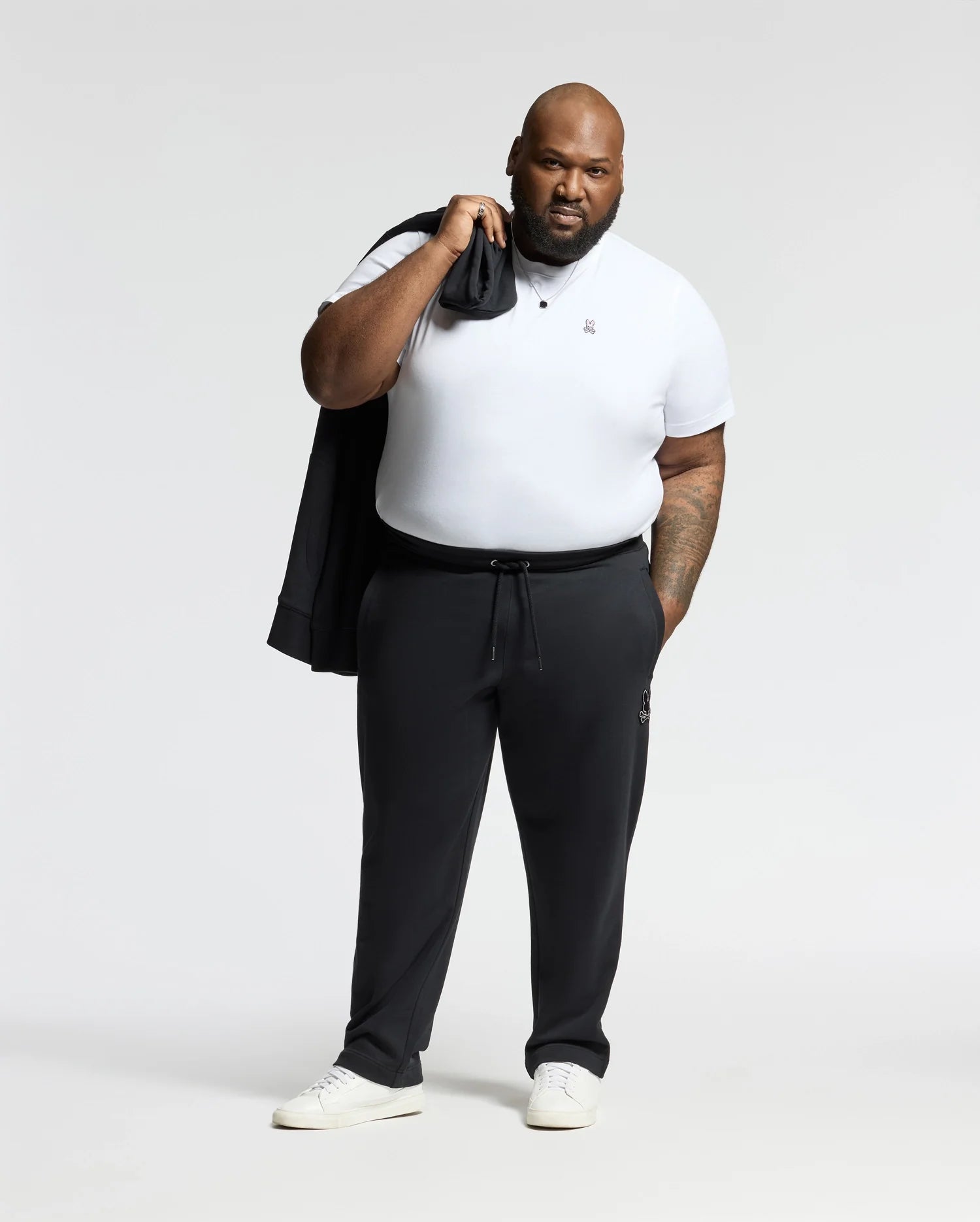 A man with a neutral expression and beard stands against a plain background, holding a jacket over his shoulder. He is dressed in a white t-shirt, BIG AND TALL ALEXANDER SWEATPANT - B9P155E200 by Psycho Bunny, and white sneakers.