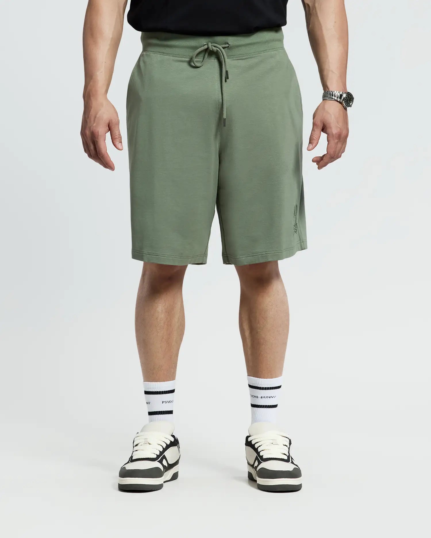 A person stands in Psycho Bunny's olive green MENS BIG AND TALL LEVANZO FRENCH TERRY SWEATSHORTS (B9R578F200) with a drawstring, paired with a black shirt, white sneakers featuring black stripes, and matching socks against a plain white background.
