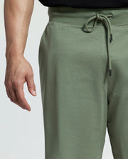 A person is wearing relaxed-fit green drawstring pants in stretch cotton jersey paired with a black shirt. The Psycho Bunny Men's Big and Tall Levanzo French Terry Sweatshorts are styled with a neatly tied drawstring, as their left hand rests naturally against a plain white background.