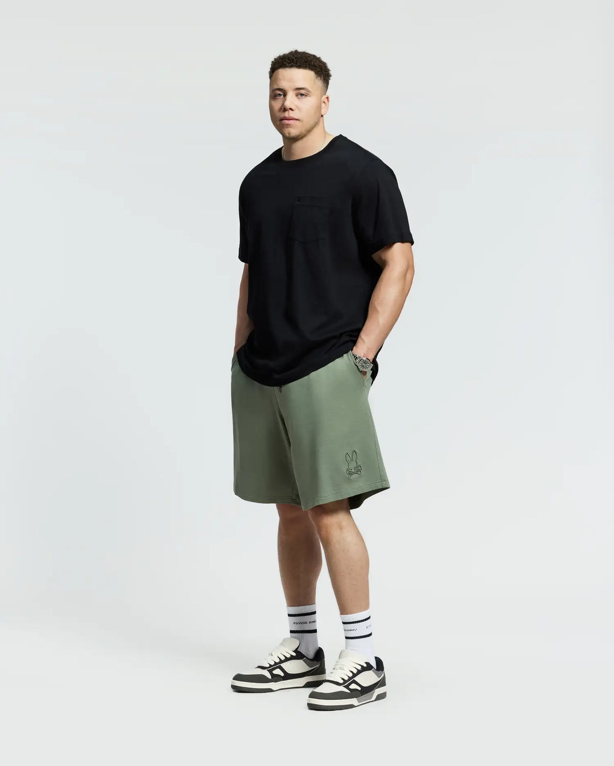 Against a plain backdrop, someone showcases effortless style wearing a black t-shirt with olive green Psycho Bunny Levanzo French Terry Sweatshorts. Paired with white socks and black-and-white sneakers, they stand facing forward with hands in pockets.
