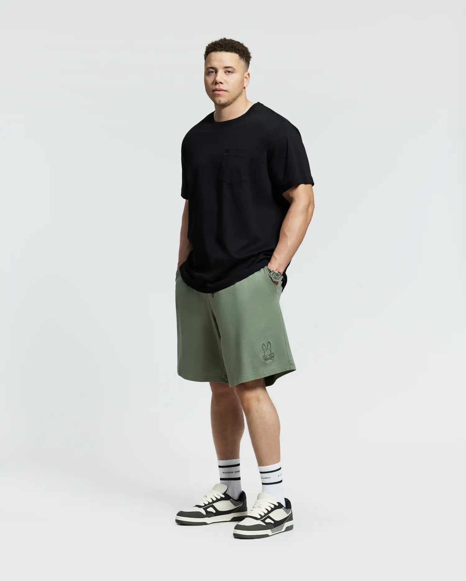 Against a plain backdrop, someone showcases effortless style wearing a black t-shirt with olive green Psycho Bunny Levanzo French Terry Sweatshorts. Paired with white socks and black-and-white sneakers, they stand facing forward with hands in pockets.
