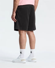 A person wearing Psycho Bunny's Mens Big and Tall Willis Stretch Tencel Shorts in black and a pink shirt with a subtle bunny pattern stands with their back to the viewer. They're sporting white socks with a black logo and white sneakers against a plain white background.