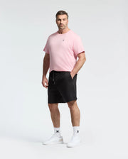 A man stands against a plain background, wearing a subtle bunny pattern pink T-shirt, Psycho Bunny shorts (MENS BIG AND TALL WILLIS STRETCH TENCEL SHORT - B9R584F200), white socks, and white sneakers. His hands rest at his sides with a neutral expression.