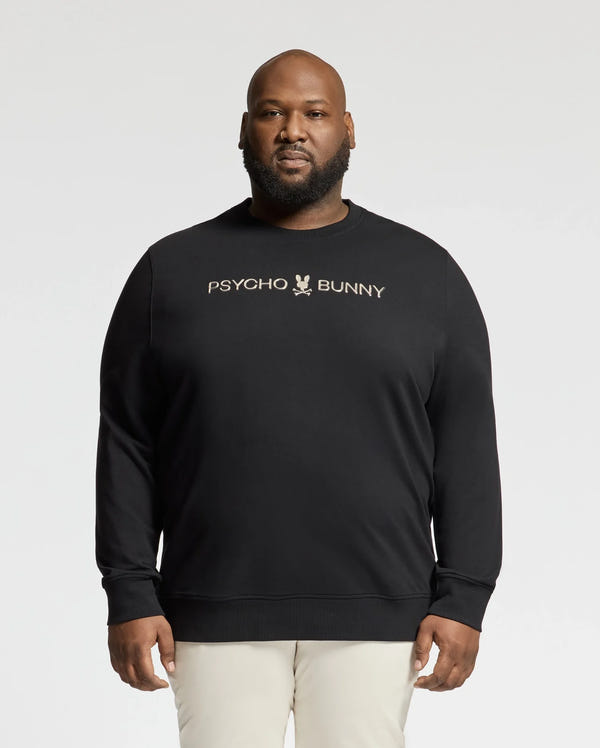 Someone in a black Psycho Bunny Big and Tall Lavata French Terry Sweatshirt (B9S159E200), featuring the logo and text on the front, stands against a plain background, stylishly paired with light-colored pants.