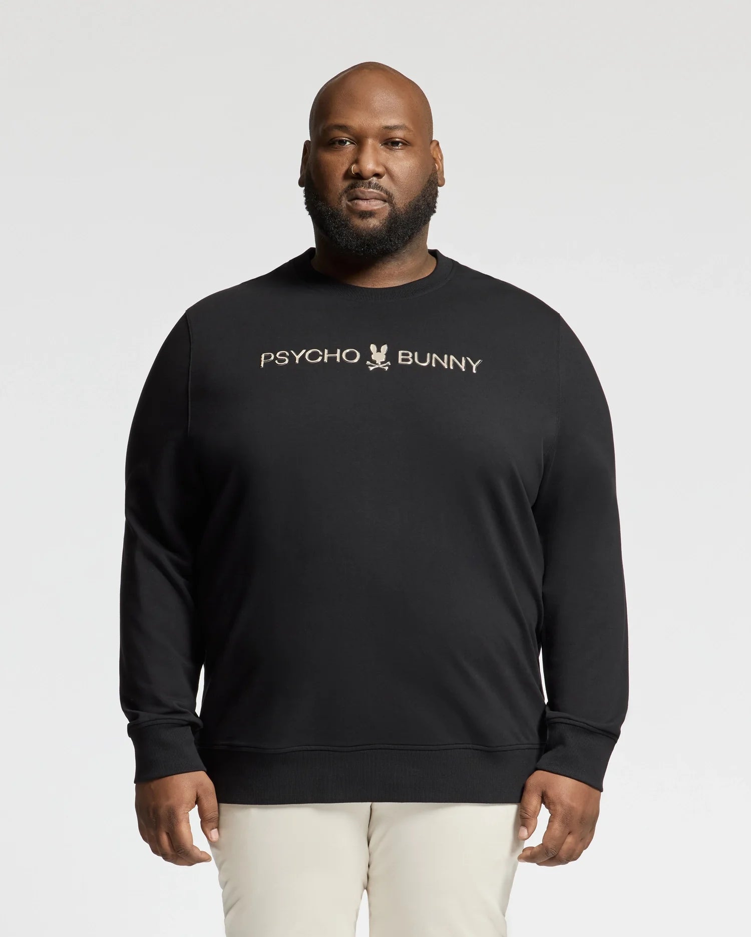 Someone in a black Psycho Bunny Big and Tall Lavata French Terry Sweatshirt (B9S159E200), featuring the logo and text on the front, stands against a plain background, stylishly paired with light-colored pants.