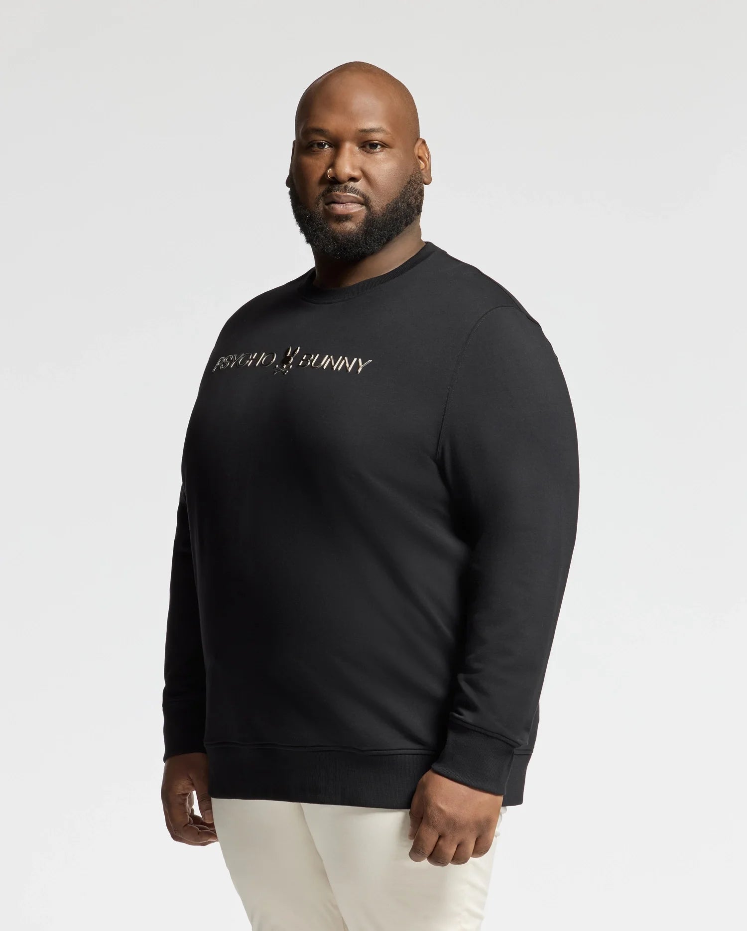 A person wearing a BIG AND TALL LAVATA FRENCH TERRY SWEATSHIRT by Psycho Bunny in black with white lettering and white pants stands against a plain white background.