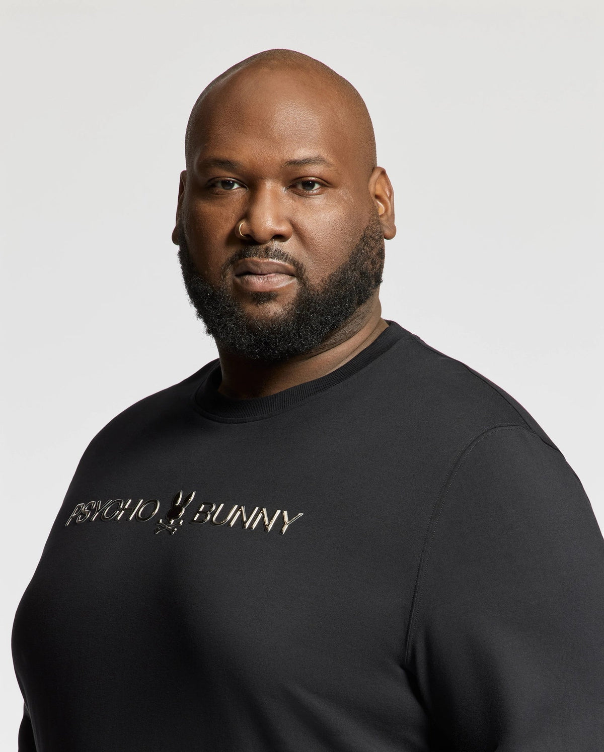 A man with a beard is wearing the BIG AND TALL LAVATA FRENCH TERRY SWEATSHIRT by Psycho Bunny, featuring "Psycho Bunny" in metallic lettering. He stands against a plain light background, gazing intently at the camera.