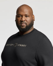 A man with a beard is wearing the BIG AND TALL LAVATA FRENCH TERRY SWEATSHIRT by Psycho Bunny, featuring "Psycho Bunny" in metallic lettering. He stands against a plain light background, gazing intently at the camera.