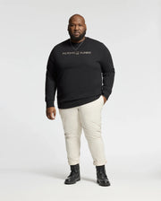 A person stands against a neutral background wearing the BIG AND TALL LAVATA FRENCH TERRY SWEATSHIRT - B9S159E200 by Psycho Bunny, paired with beige pants and black boots. The comfortable sweatshirt fits well as the figure faces forward with a calm expression.