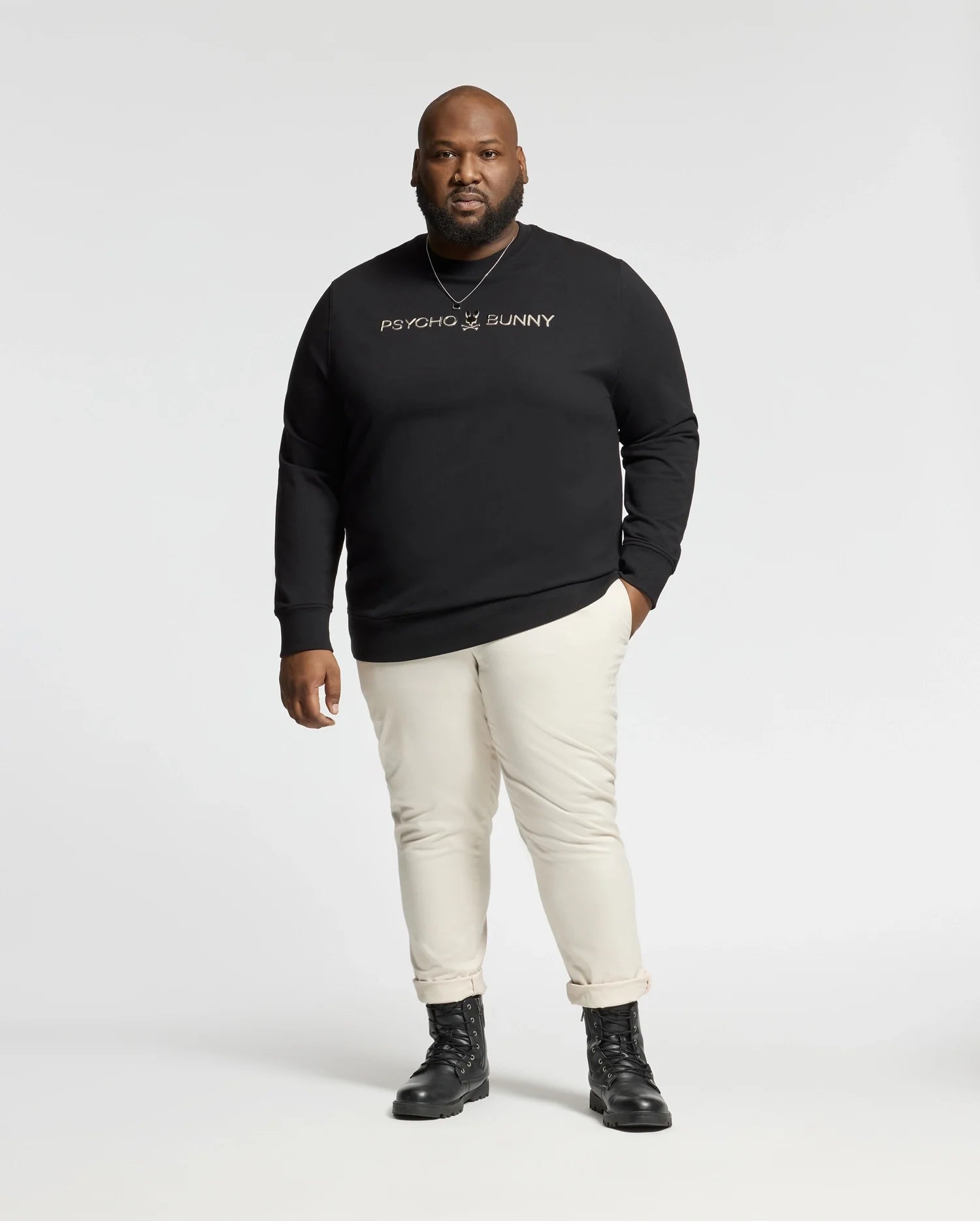 A person stands against a neutral background wearing the BIG AND TALL LAVATA FRENCH TERRY SWEATSHIRT - B9S159E200 by Psycho Bunny, paired with beige pants and black boots. The comfortable sweatshirt fits well as the figure faces forward with a calm expression.